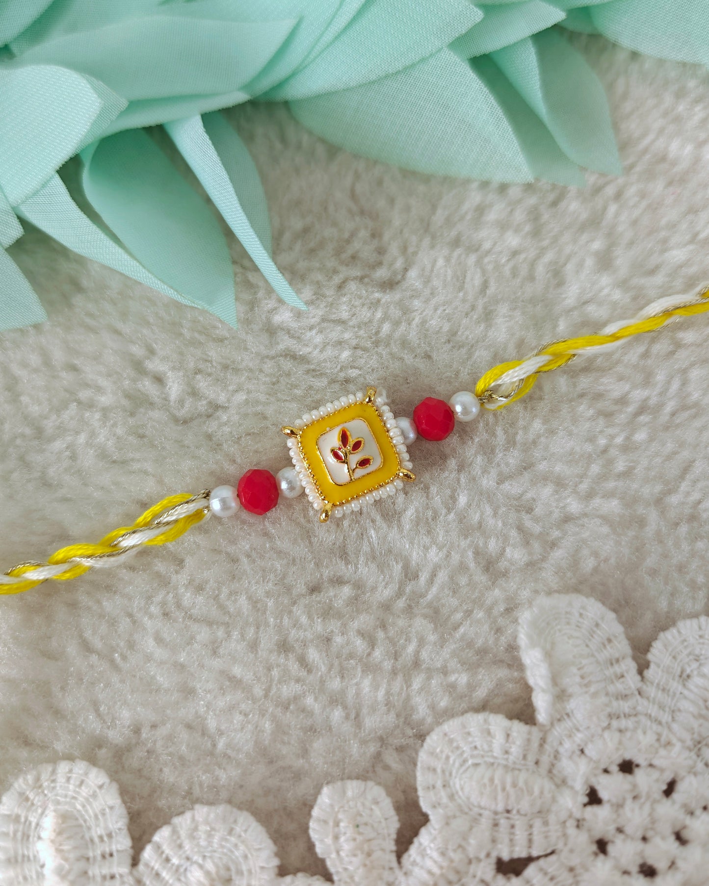 Yellow square floral rakhi with pearl border, red bead accents and twisted threads on soft background